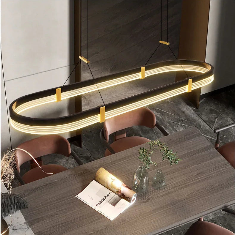 An oval-shaped modern linear LED pendant light with a black frame and warm yellow light, hanging over a dark wood dining table with brown chairs.