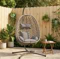 Vela Hanging Egg Chair in natural rattan with grey cushions on a stand, garden patio setting