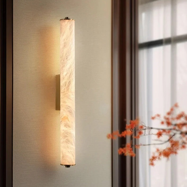 Calacatta natural marble cylinder wall light in brushed gold glowing warm white beside a living room window