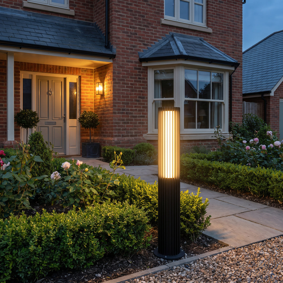 Lucevia fluted black outdoor bollard light glowing on a garden path in front of a brick house at dusk - IP65 pathway lighting